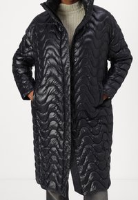 Abrigo negro acolchado con un patrón ondulado, cierre frontal con cremallera, cuello alto y bolsillos laterales. Textura lisa y brillante, diseño hasta la rodilla.