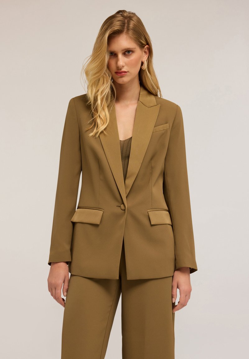 Olivově zelený na míru šitý blazer s klopami, dvěma předními kapsami a knoflíkovým zapínáním, spárovaný s odpovídajícími kalhotami širokého střihu. Hladká textilie.