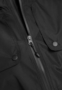 Veste en tissu noir avec fermeture éclair centrale argentée et deux poches à rabat boutonnées de chaque côté.