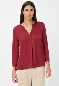 Mujer con cabello largo y oscuro, vistiendo una blusa roja de manga tres cuartos y pantalones claros con estampado, de pie contra un fondo blanco liso.