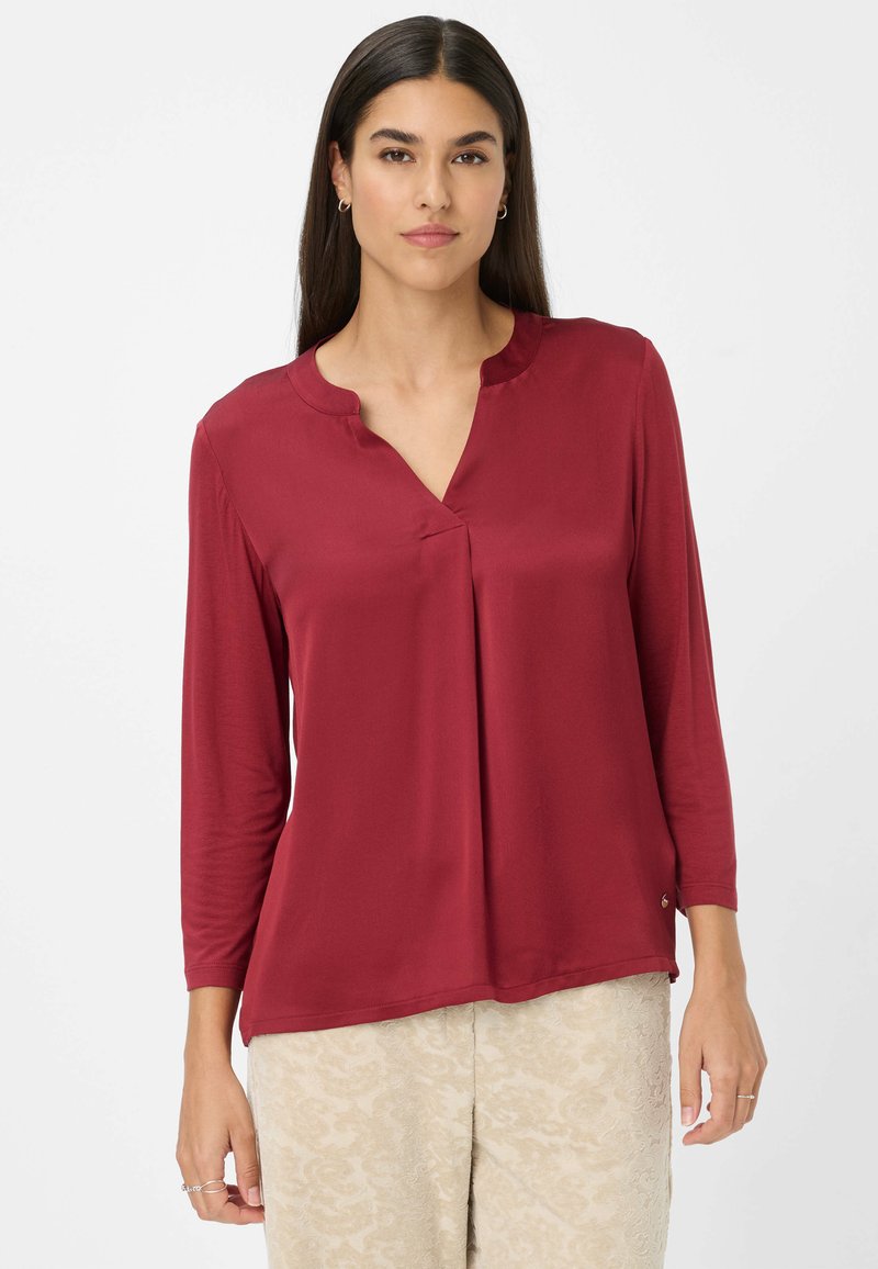 Mujer con cabello largo y oscuro, vistiendo una blusa roja de manga tres cuartos y pantalones claros con estampado, de pie contra un fondo blanco liso.