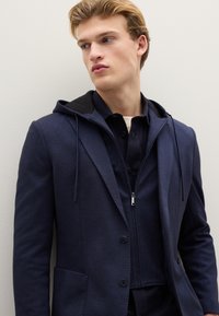 Blazer sportivo blu navy con cappuccio, caratterizzato da una texture sottile, tasche frontali e uno strato interno con zip. Chiusura con bottoni con dettagli neri.