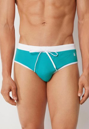 Torace maschile con slip da bagno turchese con fascia bianca e coulisse, in piedi su sfondo uniforme.