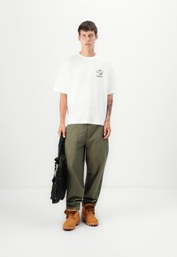 White cotton t-shirt with black logo, loose olive green pants, and tan lace-up boots. Black bag held in left hand. Simple, casual style.