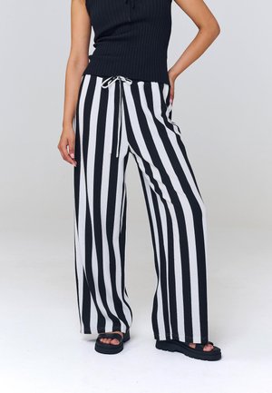 Henkilö pukeutuneena mustaan hihattomaan toppiin ja leveälahkeisiin mustavalkoraidallisiin housuihin sekä mustiin korollisiin sandaaleihin.