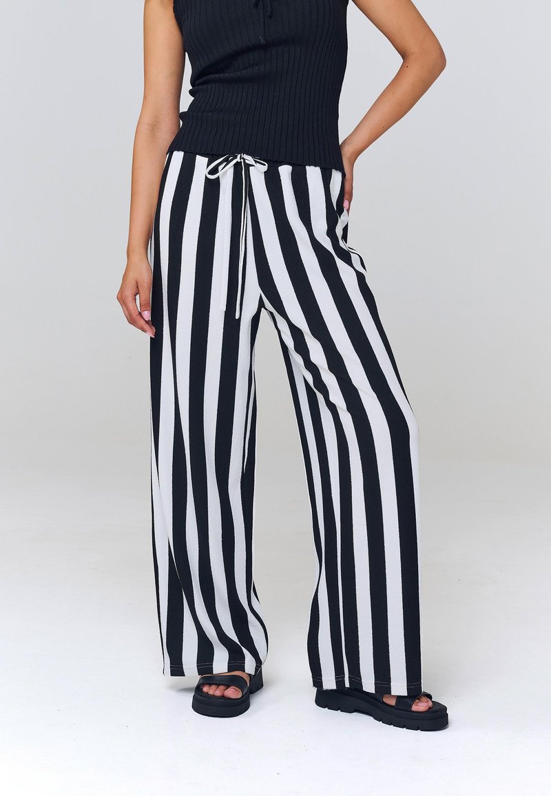 Personne portant un haut noir sans manches et un pantalon large rayé vertical noir et blanc avec des sandales compensées noires.