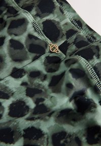 Gros plan sur un tissu imprimé léopard vert et noir avec des coutures cousues et un petit détail de logo rond doré.