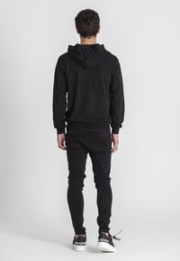 Crna hoodie s prednjim džepom i podesivom kapuljačom, uparen s uskim crnim trapericama i bijelim tenisicama s crnim akcentima.