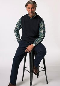 Námořnický svetř v krku přes košili s kostkovaným vzorem v zelené a modré barvě, v kombinaci s tmavými kalhotami a hnědými koženými botami, sedící na černém stoličku.