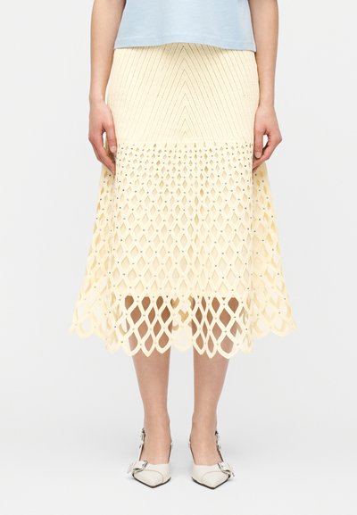 Femme portant un haut bleu clair avec une jupe midi couleur crème présentant un motif en treillis texturé et des escarpins blancs à bout pointu avec des boucles.