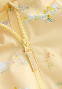 Kollane, kerge jope, millel on esikinnituseta tõmblukk ja pastelsed loomade ning lillede mustrid siledal kangapinnal.