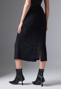 Schwarze Midirock mit einem subtilen strukturierten Muster, verziert mit kleinen weißen Akzenten. Mit einem Seitenschlitz für Bewegungsfreiheit. Schwarze Knöchelstiefel.