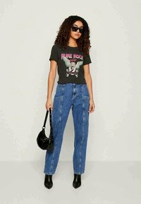 Camiseta gráfica preta com uma estampa ousada, jeans de cintura alta azuis, botas pretas de cano curto e uma pequena bolsa preta, apresentando um ajuste e textura casuais.