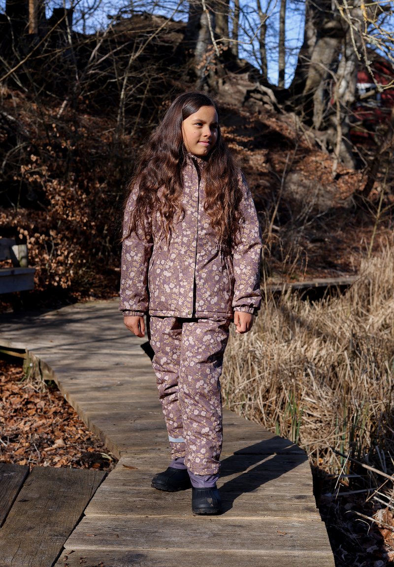 Paarse jas en broek met bloemmotief, gemaakt van zacht materiaal. Voorzien van elastische boorden en zwarte waterdichte laarzen.