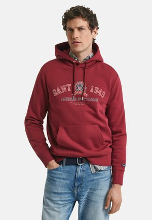 Roter Baumwollhoodie mit einer vorderen Kängurutasche, weißen Logos und Zugbändern. Über einem Hemd mit Kragen getragen und kombiniert mit blauen Jeans.