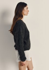 Frau mit langen braunen Haaren, die einen dunklen, flauschigen Pullover und hellgraue Shorts trägt und seitlich vor einem einfarbigen hellen Hintergrund steht.