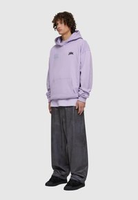 Felpa viola con tasca frontale e testo grafico, abbinata a pantaloni grigi larghi. L'outfit è completato da sneakers nere.