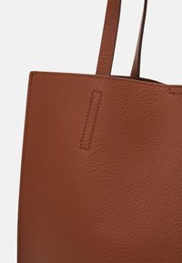 Sac fourre-tout en cuir marron avec une surface texturée, doté de deux poignées robustes et d'un design minimaliste avec un détail de couture subtil.