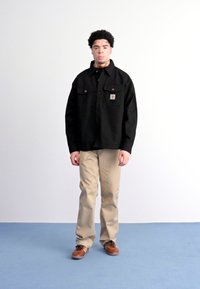 Sort, knappet skjortejakke med to brystlommer, båret over en sort skjorte, beige bukser og brune sko på et blåt gulv.