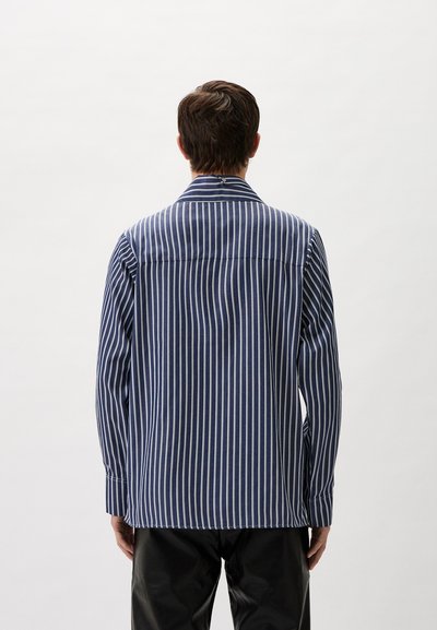 Herren Langarmhemd mit vertikalen blau-weißen Streifen, mit Kragendesign, aus glattem Stoff und einem leicht lockeren Schnitt, getragen mit schwarzen Hosen.