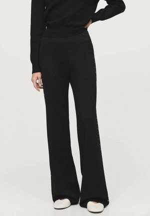 Black knit flared pants featuring side snap detailing, smooth texture, and a fitted waistband, paired with white slip-on shoes.