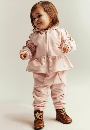 Vaaleanpunainen puuvillainen huppari, jossa on röyhelöhihat ja peplumhelma, yhdistettynä samanlaisiin röyhelöhelmaisiin housuihin ja ruskeisiin nyörikenkiin.