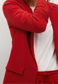 Rode blazer met gestructureerde schouders, gladde stof en een enkele voorzak, gecombineerd met een eenvoudig wit shirt eronder.