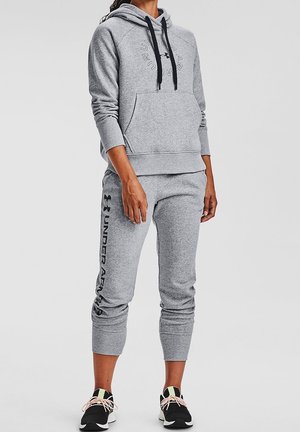 Osoba ubrana w szarą bluzę z kapturem Under Armour oraz pasujące spodnie dresowe z czarnymi sportowymi butami, stojąca na jednolitym jasnym tle.