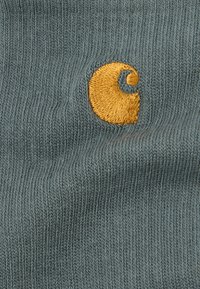 Zbliżenie na niebieską dzianinę z małym, haftowanym żółtym logo przedstawiającym zakrzywiony kształt i okrąg.