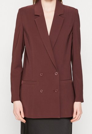 Frau trägt einen zweireihigen braunen Blazer mit Kerbkragen und vorderen Paspeltaschen, kombiniert mit einem schwarzen Rock.