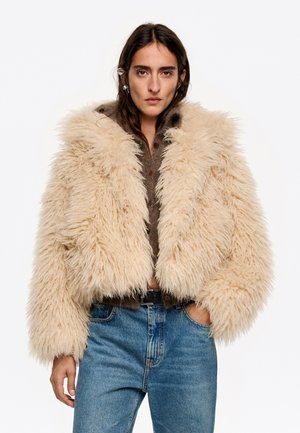 Chaqueta corta de peluche sintético beige con una textura esponjosa, cuello ancho y cierre con botones, superpuesta sobre una blusa marrón y unos vaqueros azules.