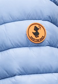 Πορτοκαλί κυκλικό σήμα με μαύρη πάπια που τραγουδά και το κείμενο «SAVE THE DUCK» σε ανοιχτό μπλε καπιτονέ ύφασμα.