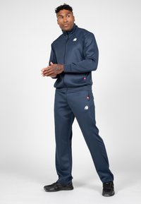 Marine tracksuit van soepel materiaal met een volledige ritsjack en broeken met elastische tailleband. Bevat logo-accenten en zijzakken. Zwarte sportschoenen.