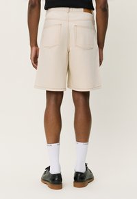 Pantalones cortos de mezclilla beige con corte recto, costuras visibles y dos bolsillos traseros. Se llevan con zapatos negros y calcetines blancos con texto.