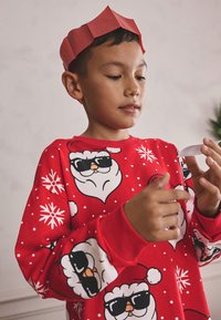 Sweat-shirt rouge à manches longues avec des pois blancs, des visages de Père Noël et des flocons de neige. Le garçon porte une couronne en papier rouge simple.