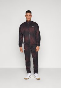 Melnās un sarkanās sporta uzvalks ar ar rāvējslēdzēju aizdari un gumijas aprocēm, kurā ir neliels zaļš logo. Savienots ar baltām sporta kurpēm un melnām biksēm.