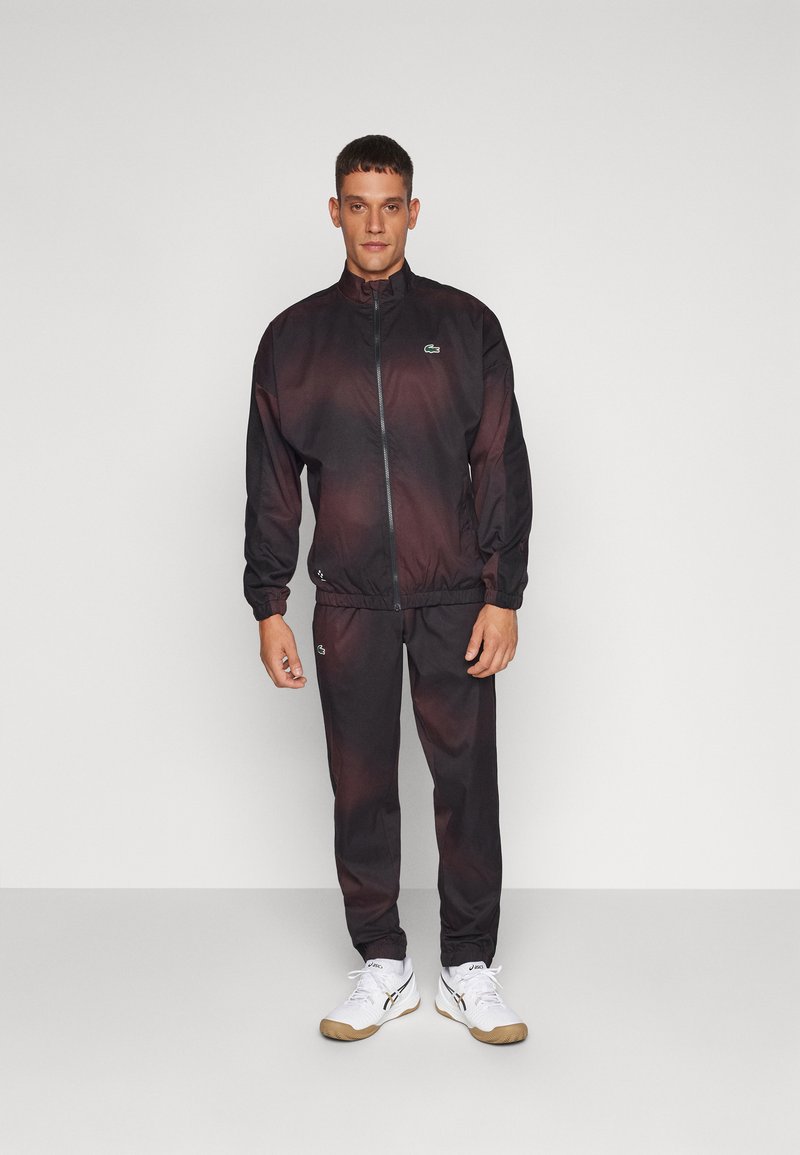 Schwarzer und roter Trainingsanzug mit Reißverschlussjacke und elastischen Bündchen, versehen mit einem kleinen grünen Logo. Kombiniert mit weißen Sneakers und schwarzen Hosen.