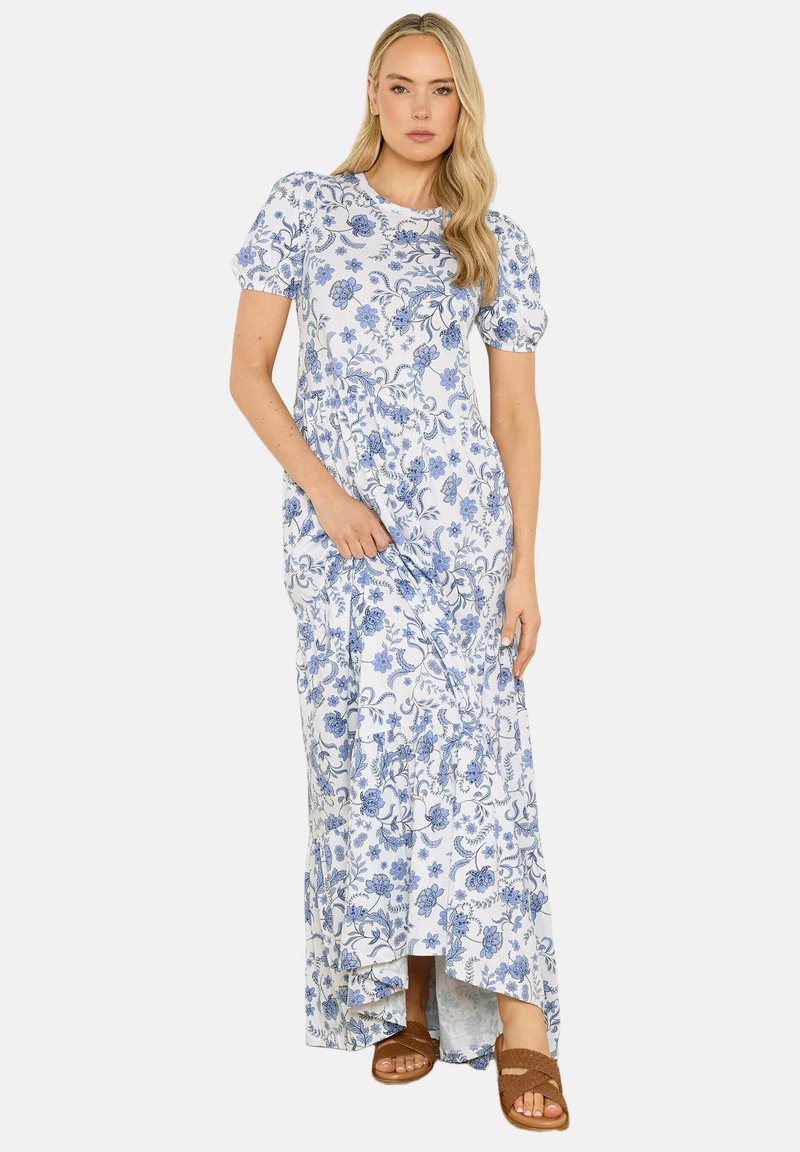 Frau in einem weißen Maxikleid mit blauem Blumenmuster, kurzen Puffärmeln und braunen Sandalen, stehend vor einem einfarbigen Hintergrund.