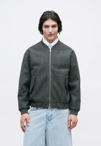 Grijze bomberjack met een gladde textuur, ritssluiting aan de voorkant en zijzakken. Gepaard met lichtblauwe denim jeans. Minimalistisch ontwerp.