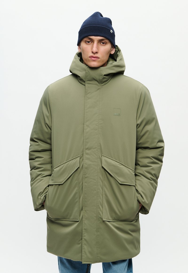 Abrigo aislante de color verde oliva con un cuello alto, dos bolsillos delanteros y un diseño sencillo. Acompañado de un gorro de lana de color azul marino.