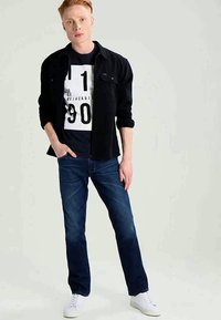 Camisa negra de botones sobre una camiseta gráfica azul marino con texto blanco, combinada con jeans azul oscuro y zapatillas blancas.