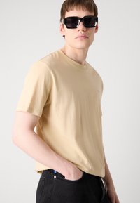 Beige bomulds T-shirt med rund halsudskæring, korte ærmer og en subtil logo detalje på brystet, parret med mørk denim jeans.