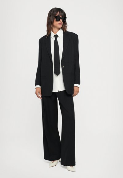 Black tailored suit with wide-leg trousers and oversized blazer, paired with a white shirt and black tie. Accented with a triangular logo.