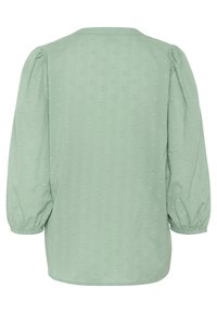 Blouse vert clair avec des manches bouffantes, encolure ronde et texture à petits cœurs sur l'ensemble. L'ourlet est légèrement courbé.