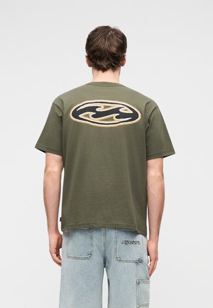 Muž v olivovo zelenej tričku s čiernym a zlatým vlnovým logom na chrbte, v svetlomodrých roztrhaných džínsoch, otočený chrbtom.