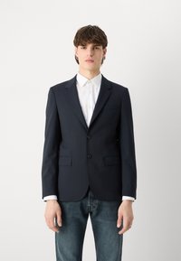 Sinine bleiser, millel on kohandatud ja sobiv lõige, kahest nupust, sisselõigatud kraest ja eesmistest taskutest. Kombineeritud valge märgiga särk.