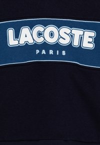 Sweatshirt bleu marine avec un grand logo blanc "LACOSTE" et le texte "PARIS" en design courbé. Présente un tissu texturé et des rayures contrastantes bleu clair.