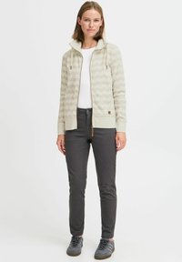 Sweat-shirt zippé beige clair avec un motif ondulé texturé, des poignets et un ourlet côtelés, associé à un pantalon slim gris foncé et des baskets grises.