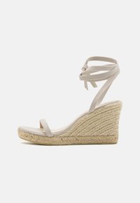 Beige kile sandal med en vevd juteplattform, minimale stropper og ankelbånd. Mykt semsket lær, buet form og åpen tå-design.