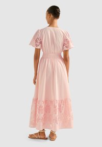 Rochie maxi din bumbac roz deschis, cu mâneci scurte și pufoase, și tiv brodat. Prezintă o talie elastică, design în straturi și sandale plate.